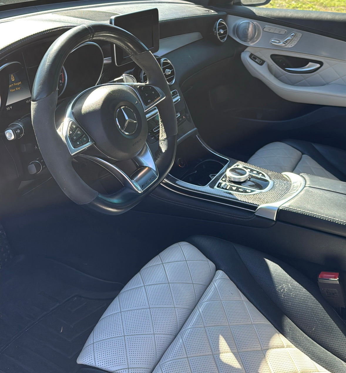 Luxury Mercedes-Benz AMG car interior featuring black leather multi-function steering wheel, quilted perforated leather seats, and carbon fiber center console with control dial.