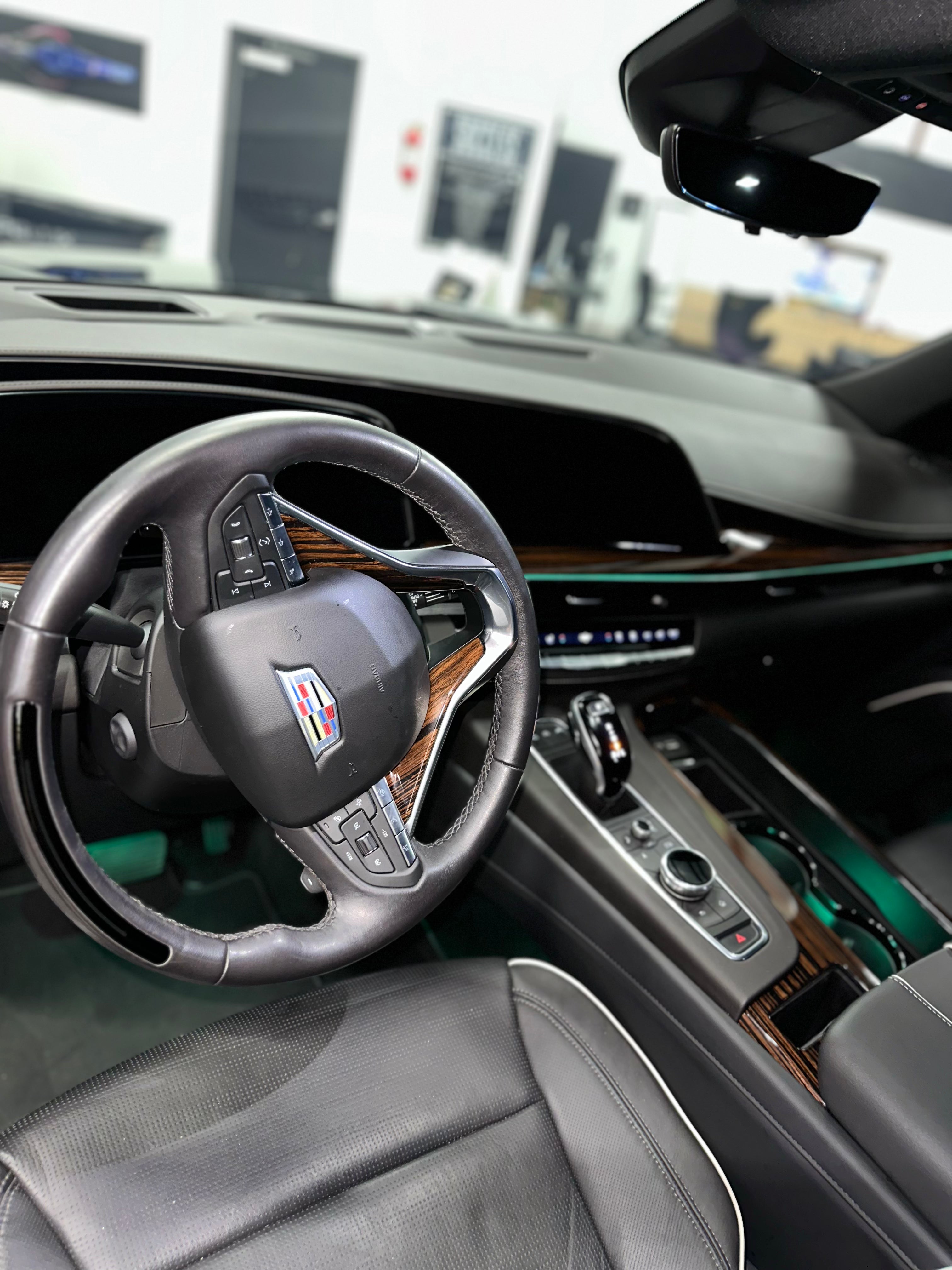 Luxury Cadillac car interior with black leather steering wheel and wood trim dashboard, premium ergonomic driver seat, and modern infotainment controls.