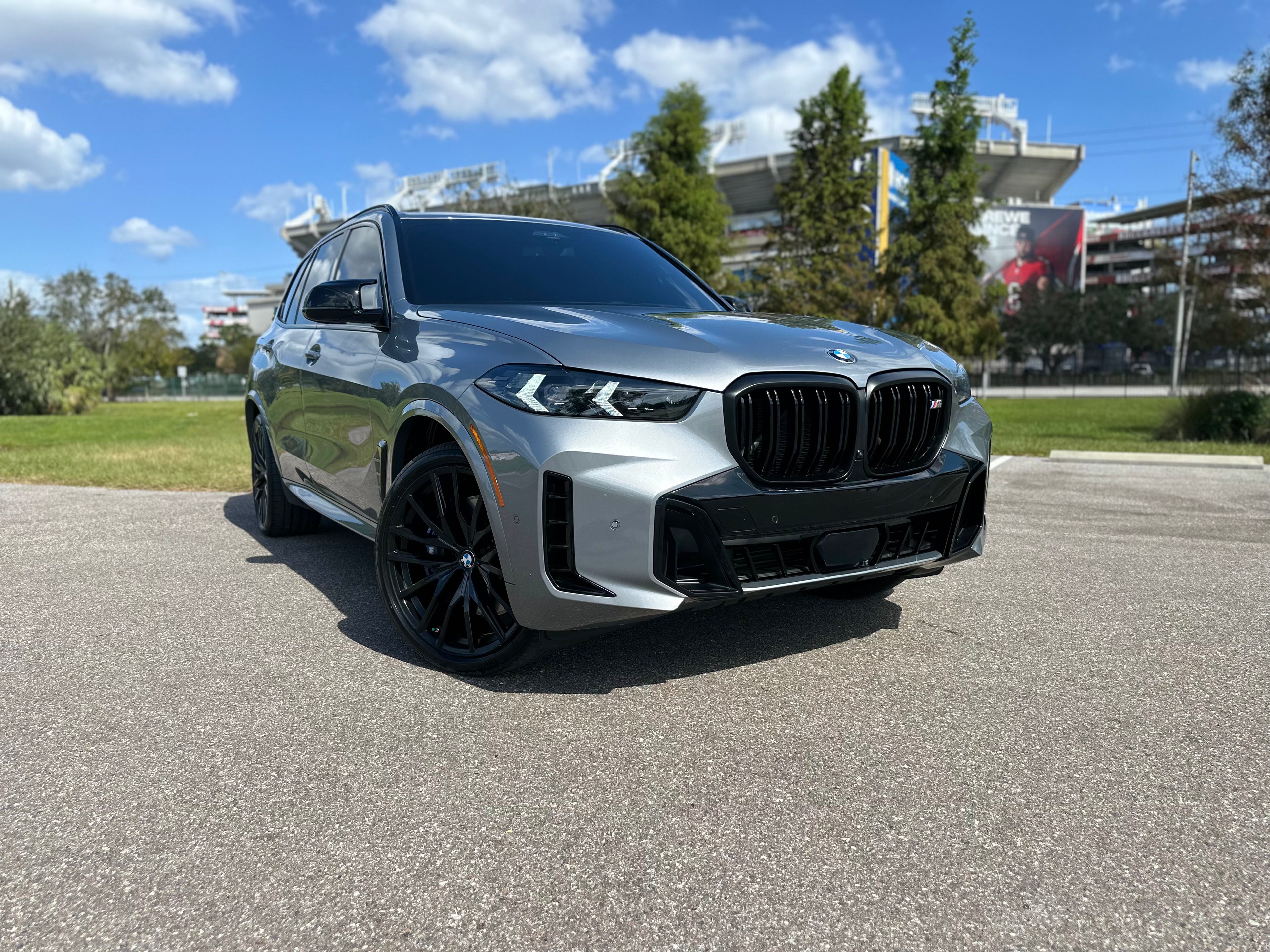 2023 BMW X5 M Sport utility vehicle in sleek metallic gray with black alloy wheels, parked outdoors under a sunny blue sky.