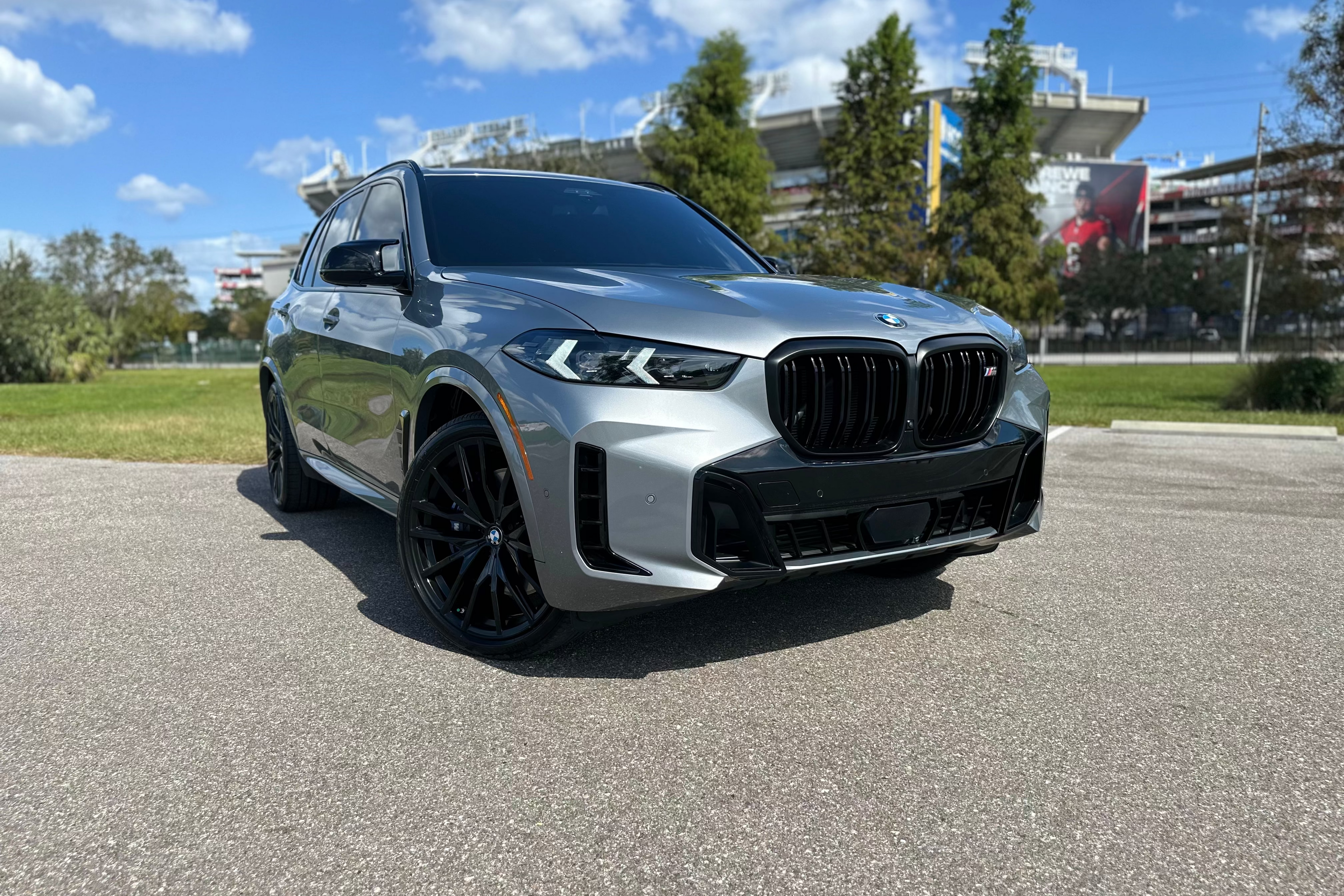 Silver BMW X5 M50i luxury SUV with black rims parked outdoors under clear blue sky, premium performance vehicle.