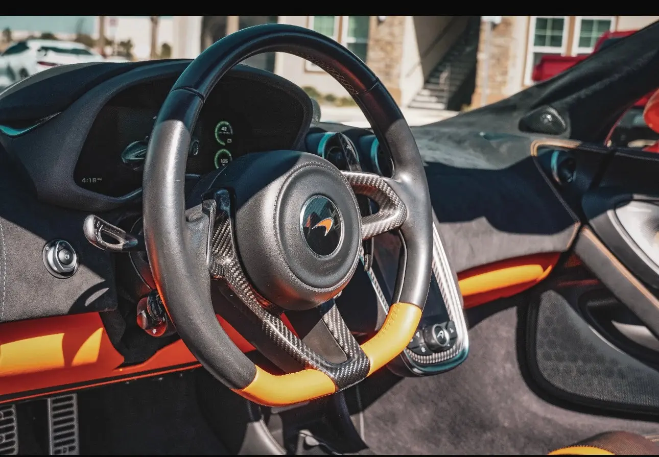 Luxury sports car steering wheel with carbon fiber and black leather, featuring orange accents and advanced dashboard controls.