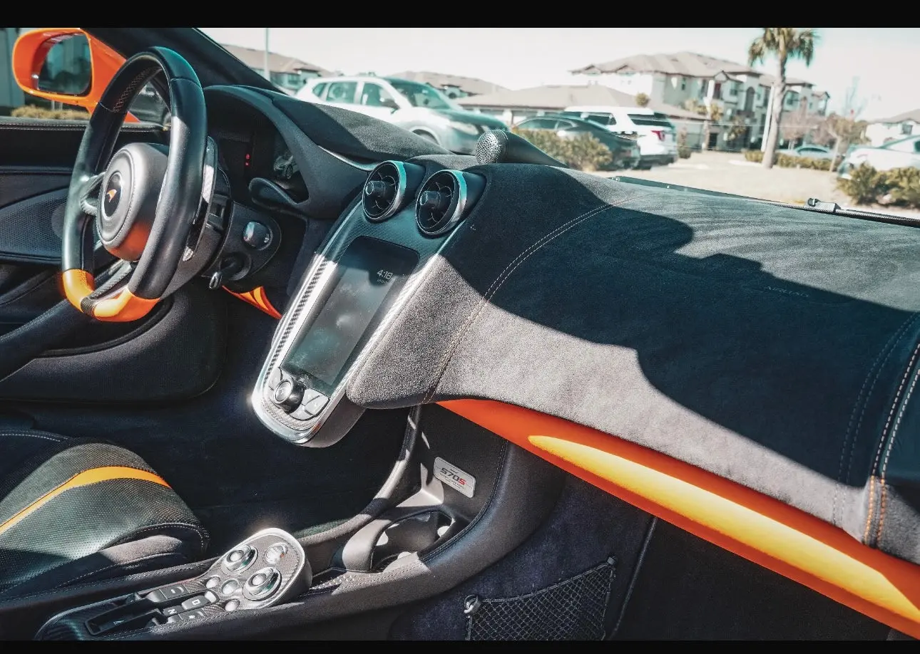 Interior of McLaren 570S sports car featuring black and orange leather seats, carbon fiber steering wheel, and touchscreen dashboard display.