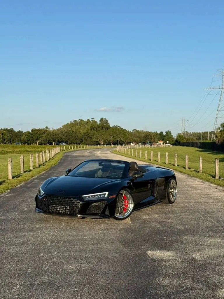 Black Audi R8 convertible sports car with red brake calipers parked on rural road under blue sky, luxury high-performance supercar.