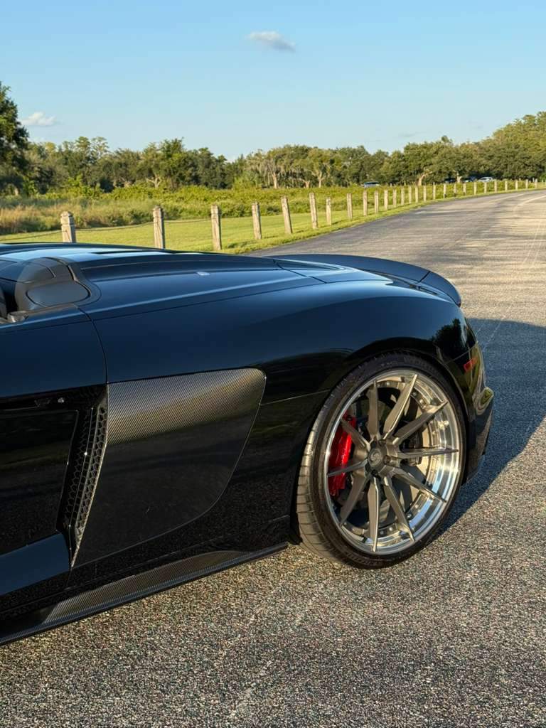 Close-up of black luxury sports car with carbon fiber side panel and polished alloy wheels featuring red brake calipers on a scenic road.