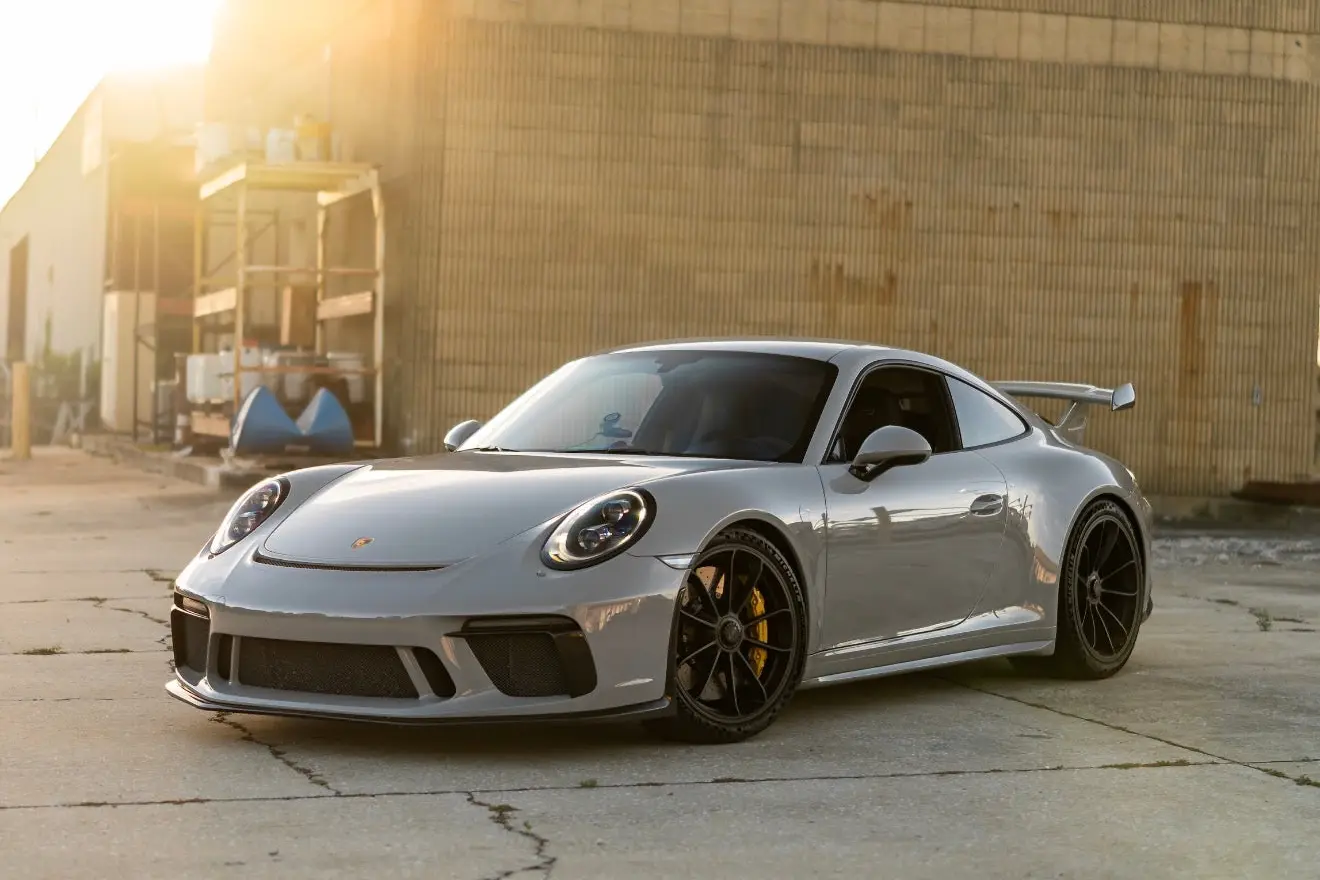 Silver Porsche 911 GT3 sports car with black alloy wheels and rear spoiler, parked on concrete near industrial building in golden hour sunlight.