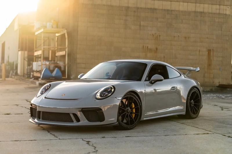 Silver Porsche 911 GT3 sports car with black alloy wheels and rear spoiler, parked on concrete near industrial building in golden hour sunlight.