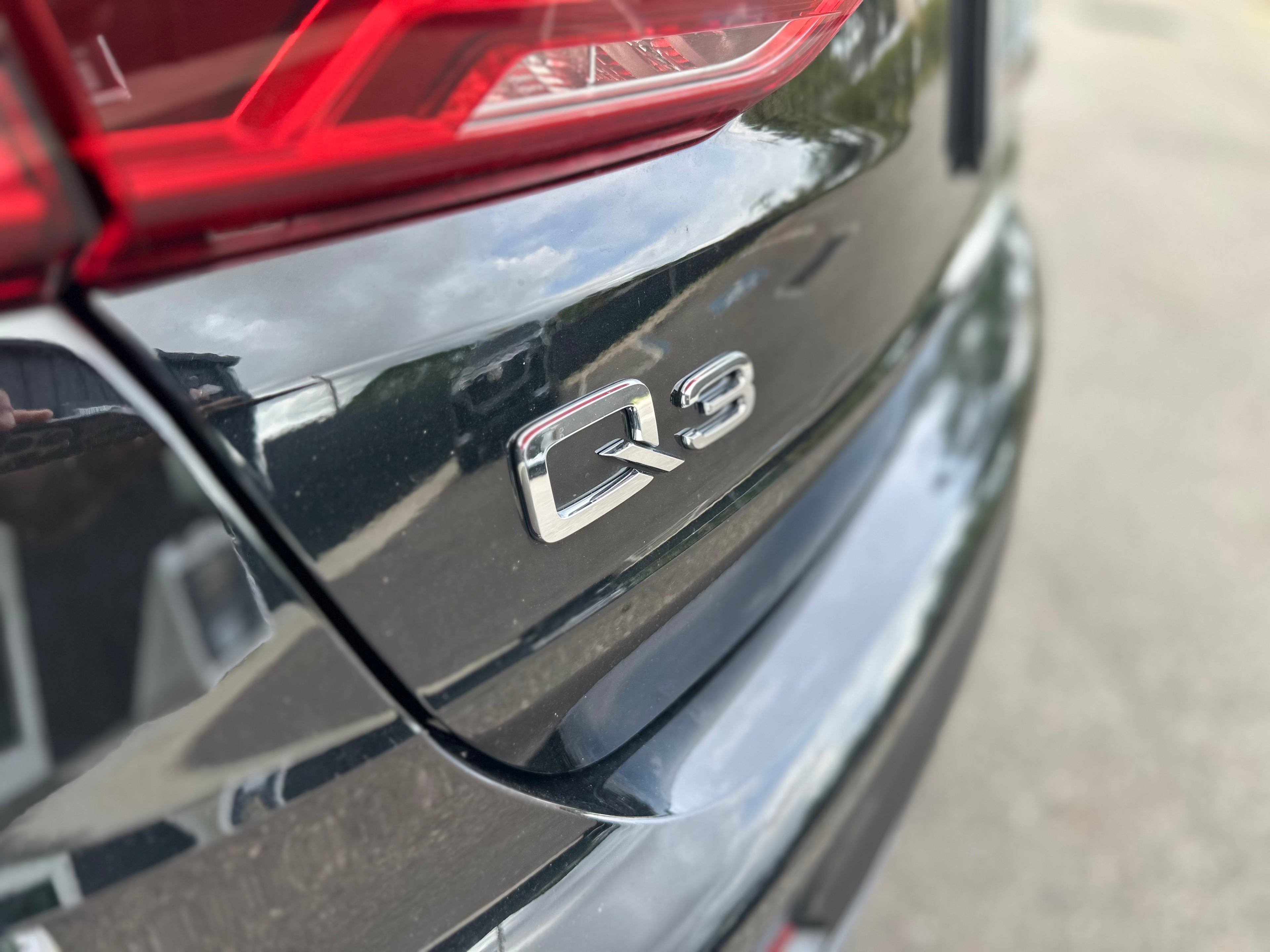 Close-up of black Audi Q3 rear badge with red taillight, featuring sleek luxury SUV design and glossy finish.
