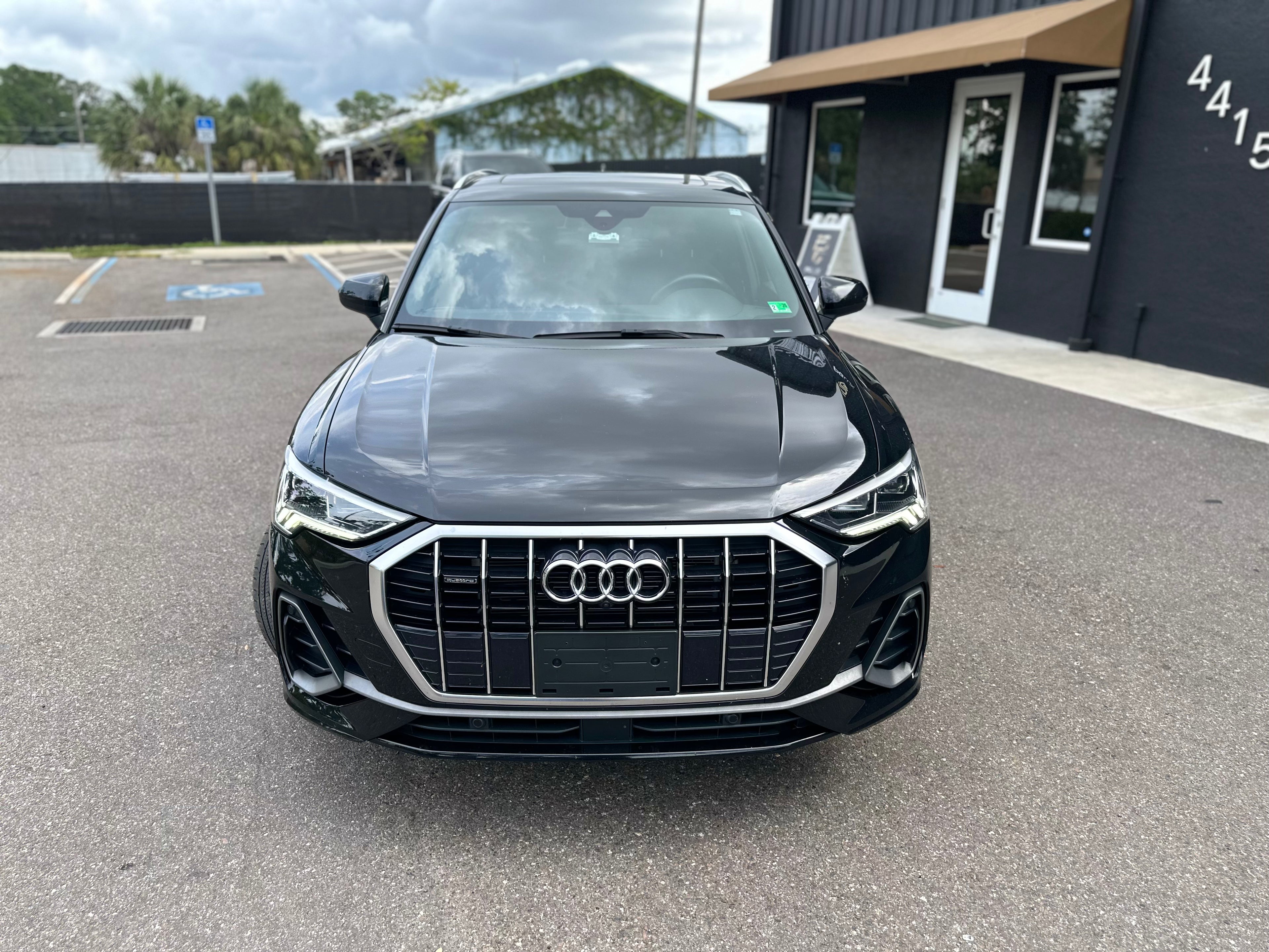 Black Audi Q3 SUV front view with LED headlights and chrome grille, parked outside showroom, luxury compact crossover car.