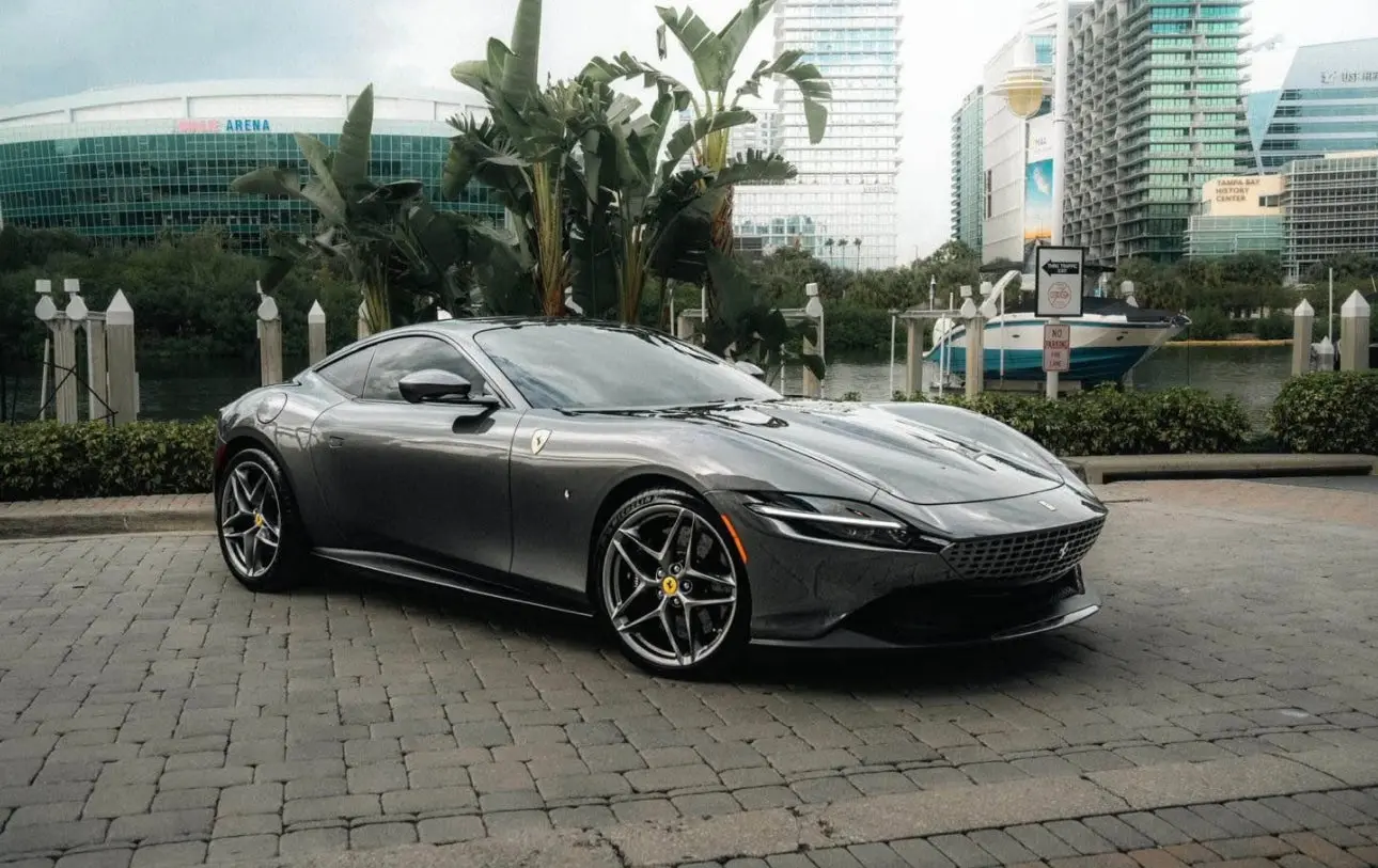 Sleek gray Ferrari Roma sports car parked on a waterfront urban street with luxury alloy wheels and aerodynamic design.