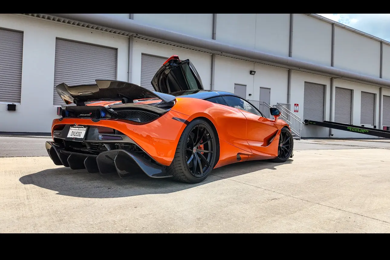 Orange McLaren supercar with black carbon fiber rear spoiler and open butterfly door parked outside industrial building, luxury sports car exterior.