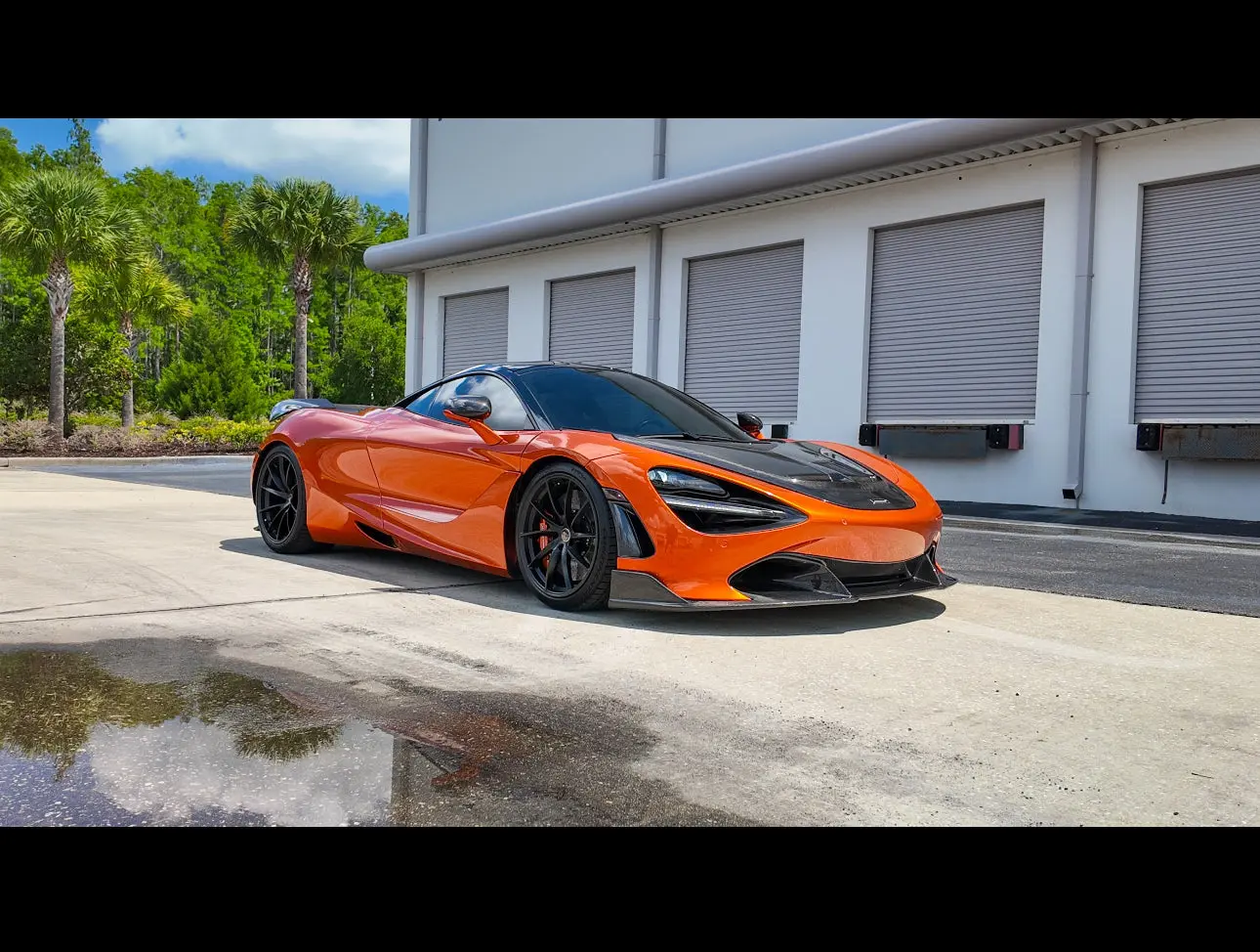 Orange McLaren 720S sports car with black carbon fiber hood and black rims parked outside a modern industrial building under clear blue sky.