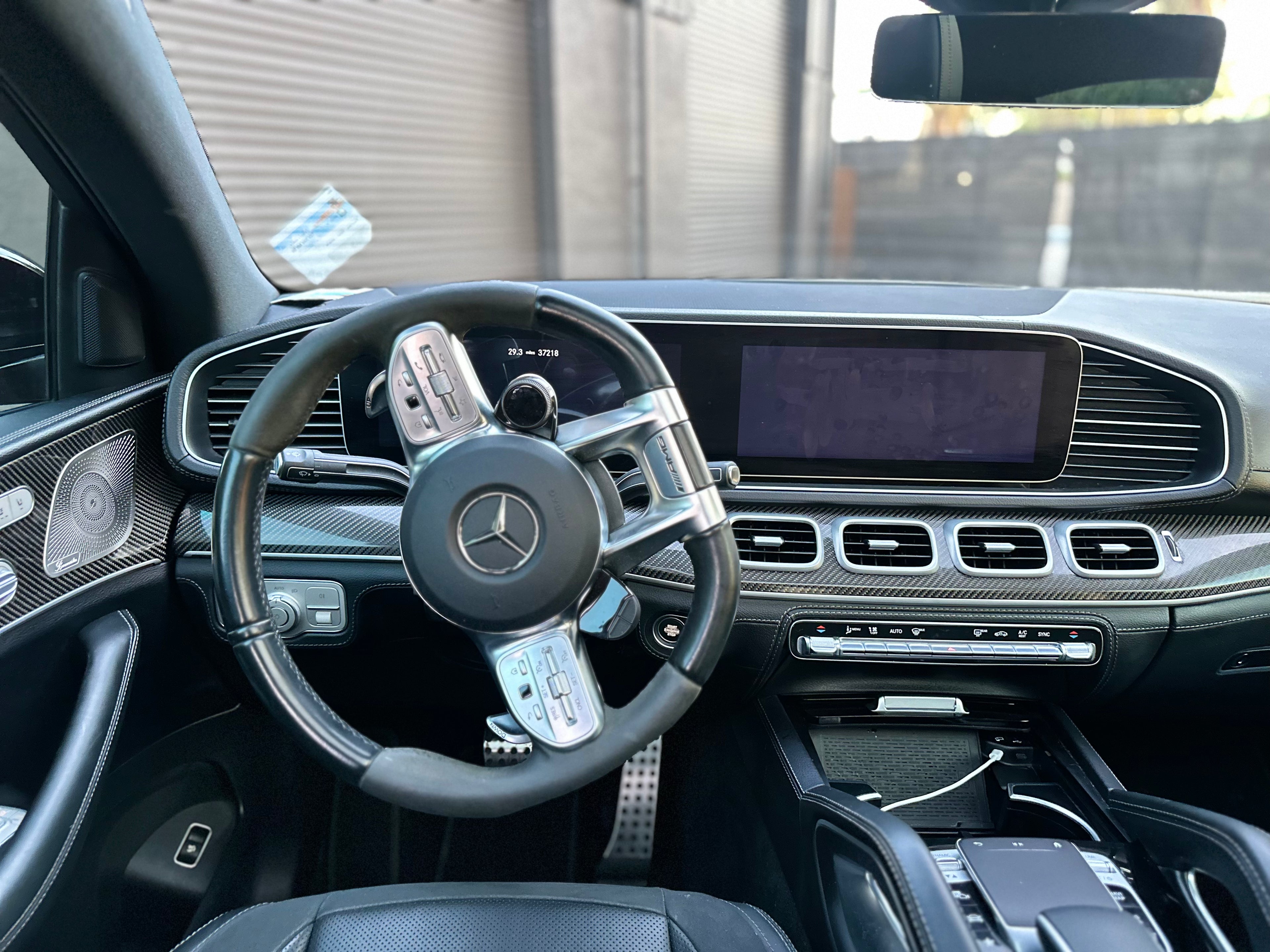 Interior view of luxury Mercedes-Benz car featuring black leather steering wheel with AMG logo, digital dashboard display, carbon fiber trim, and advanced climate control system.