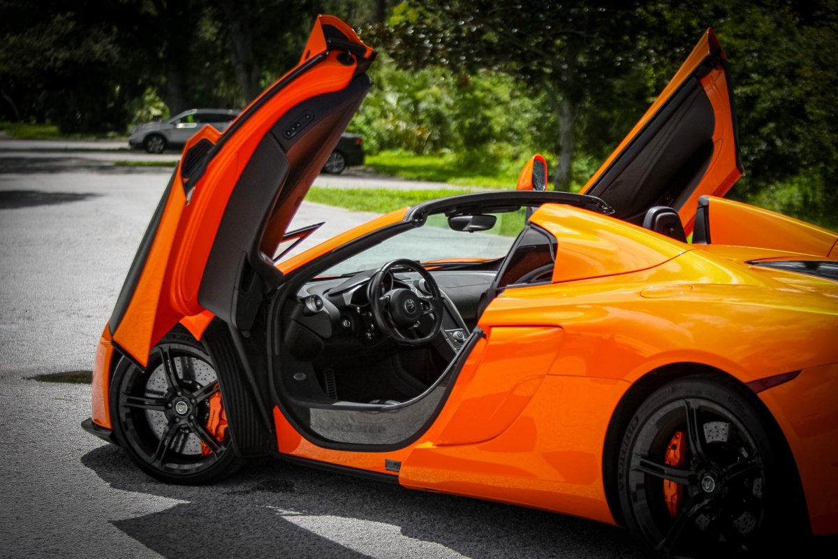 Orange luxury sports car with scissor doors open, black interior, and sleek aerodynamic design, parked outdoors on asphalt road.