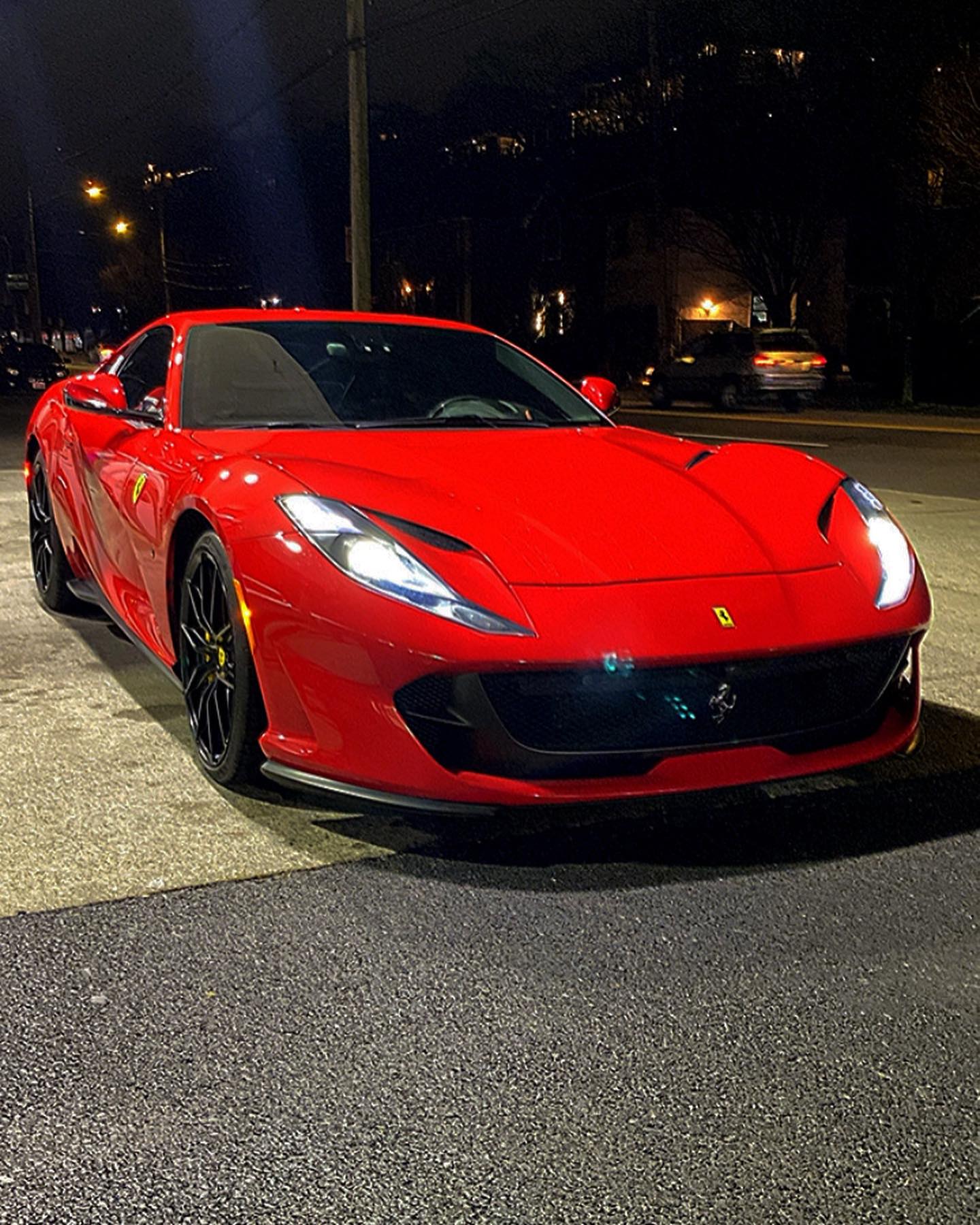 Red luxury Ferrari sports car parked on city street at night with headlights on, sleek aerodynamic design and black alloy wheels.